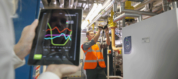 Hands holding real-time tablet data in foreground with technician adjusting pipes in background