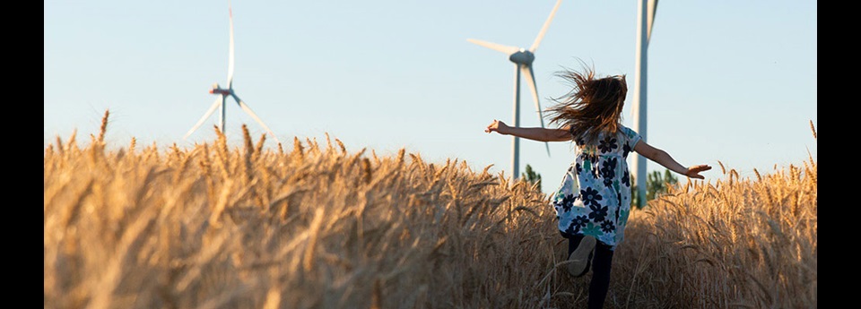 Girl running through wheat field with light blue sky and wind turbines in the distance.