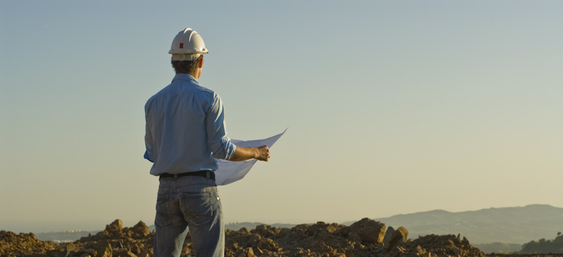 Man in hard hat holding blueprints looking over land.