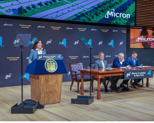 Guvenør Kathy Hochul taler ved et podium ved siden af ​​bordet med "Micron kommer til NY"-skiltet.