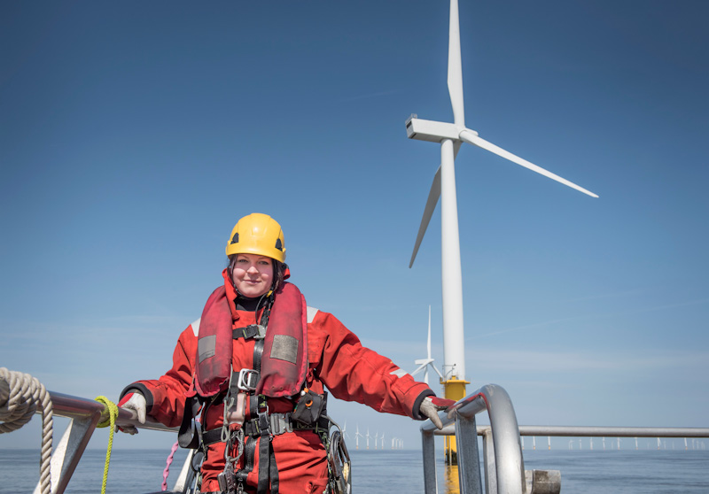 Utility worker woman in hardhat and harness in front of offshore wind turbine and blue skies.