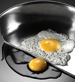 Half of a frying pan on an induction cooktop with one egg in pan and one egg directly on the cooktop. The egg in the pan is cooking and the egg on the stove is raw.