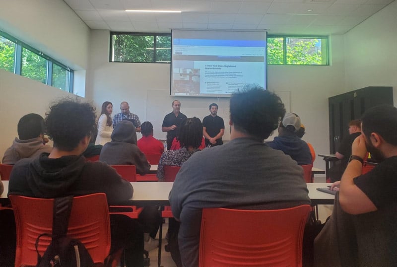 Students sitting at tables in classroom environment. Instructors at the head of the classroom in front of projector teaching curriculum focused on clean transportation.
