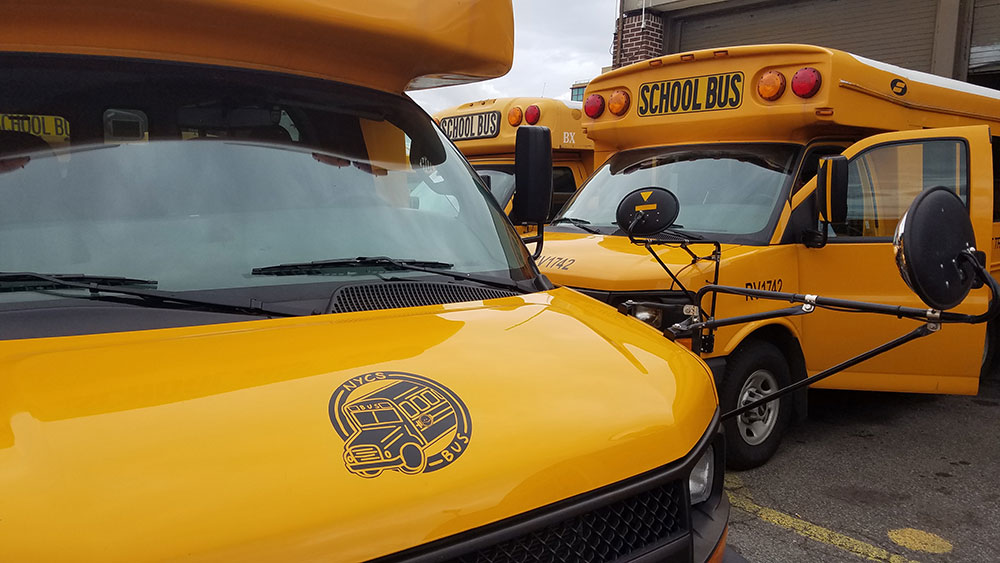 Closeup of the NYCS BUS logo on the hood of a school bus with more busses parked in background.