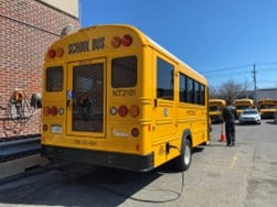 Electric school bus parked outside school bus garage attached to charging station plug.