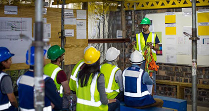 People in hard hats and safety vests sitting on benches watching a presenter holding a harness.