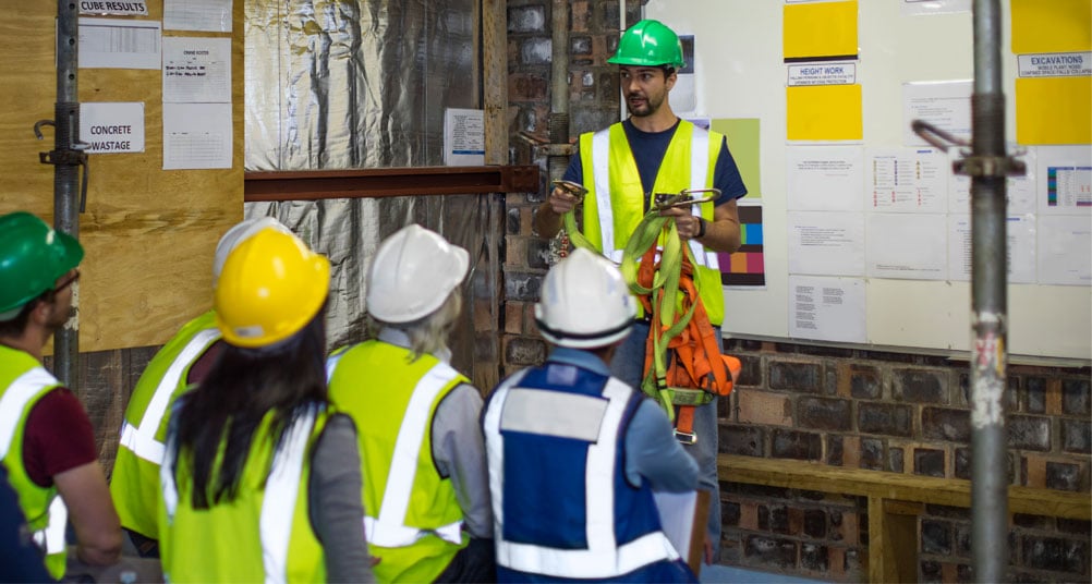 People in hard hats and safety vests sitting on benches watching a presenter holding a harness.
