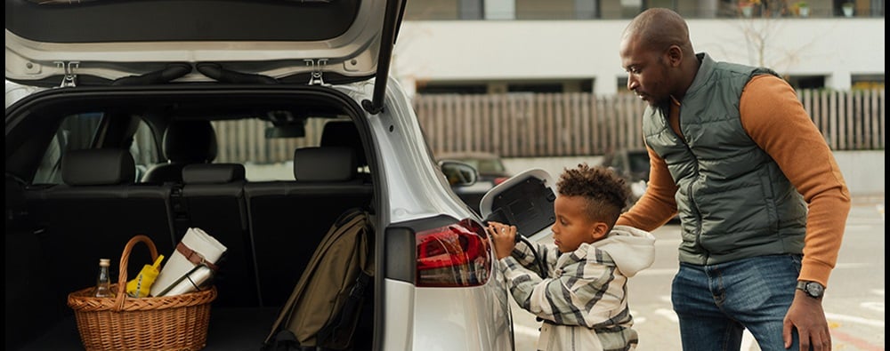 A father helping young son plug in an electric vehicle with the trunk opened in a parking lot.