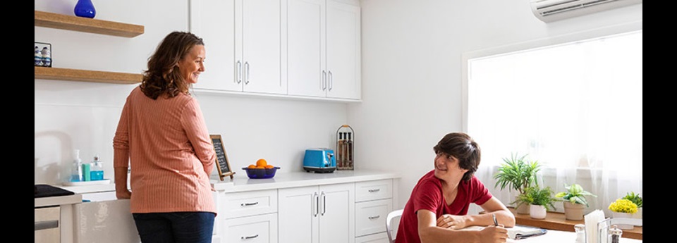 Woman at a sink smiling over her shoulder to a young man sitting at the table working on a paper.