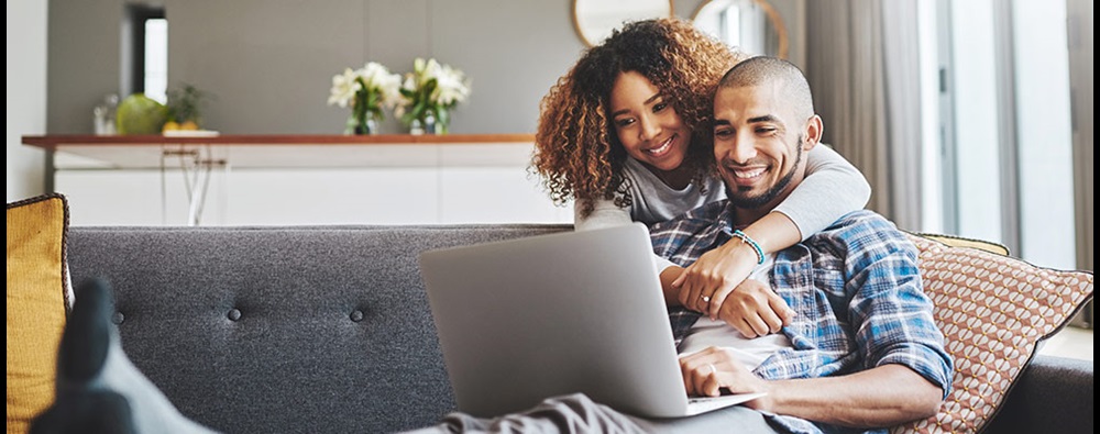 A man lounging on a couch with a woman hugging him from behind. Both are smiling and looking at a laptop.