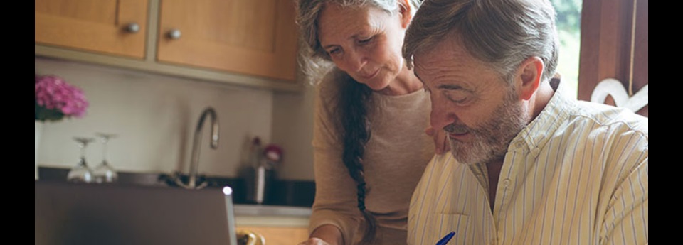 An older couple in a kitchen working on something out of frame with pen in hand and laptop on table.