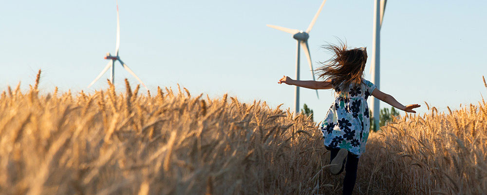 Girl running through wheat field with light blue sky and wind turbines in the distance.